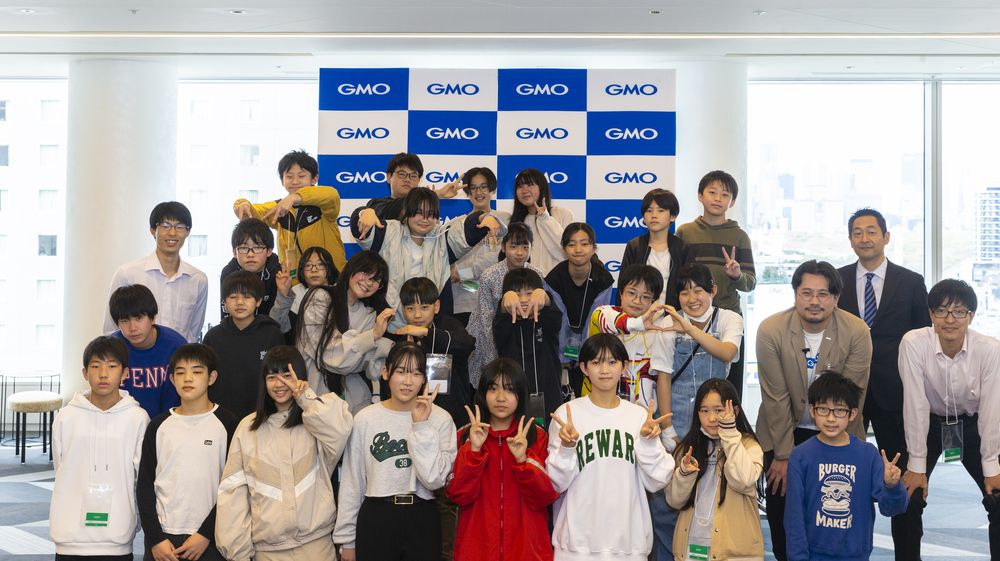 図1 写真。発表会後の集合写真。児童たちは笑顔でさまざまなピースサイン。右側にはGMOの成瀬氏、笹塚小学校の先生、渋谷区教育委員会の方々。